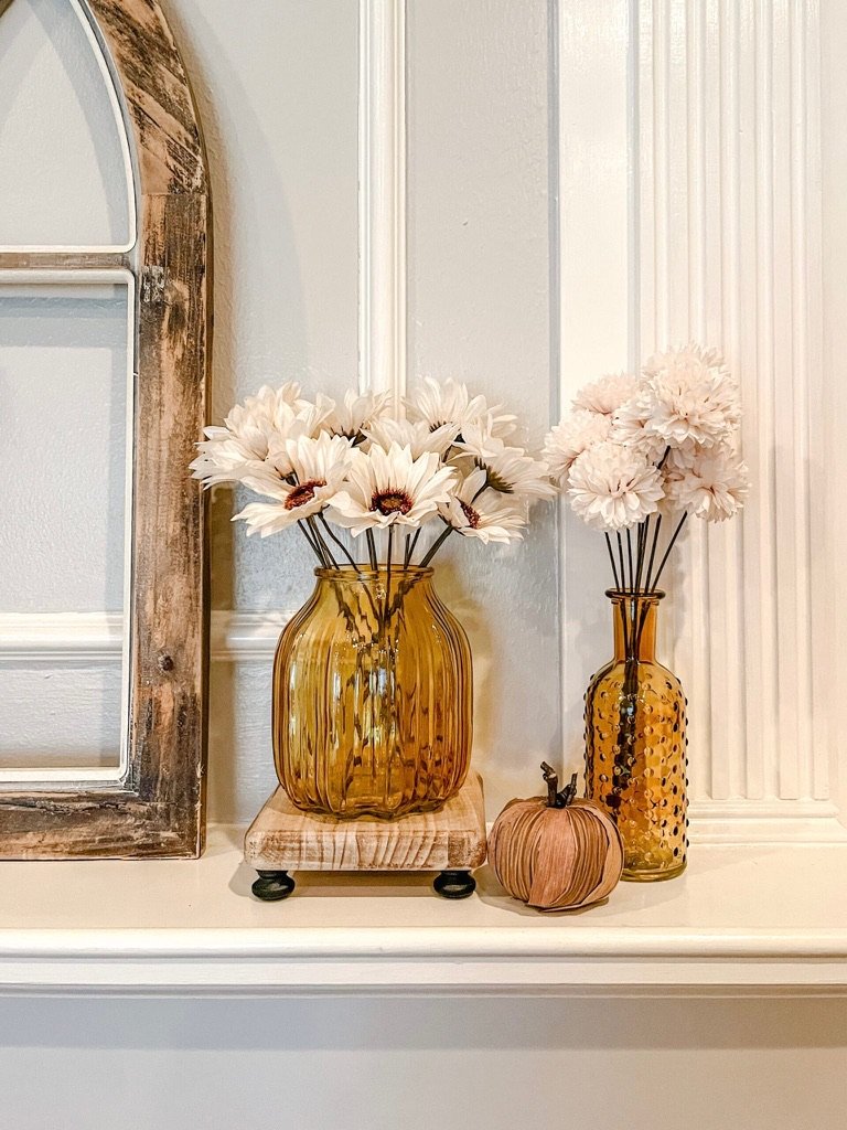 Neutral fall decor on a fireplace mantel with amber glass vases, cream florals, and a small brown pumpkin