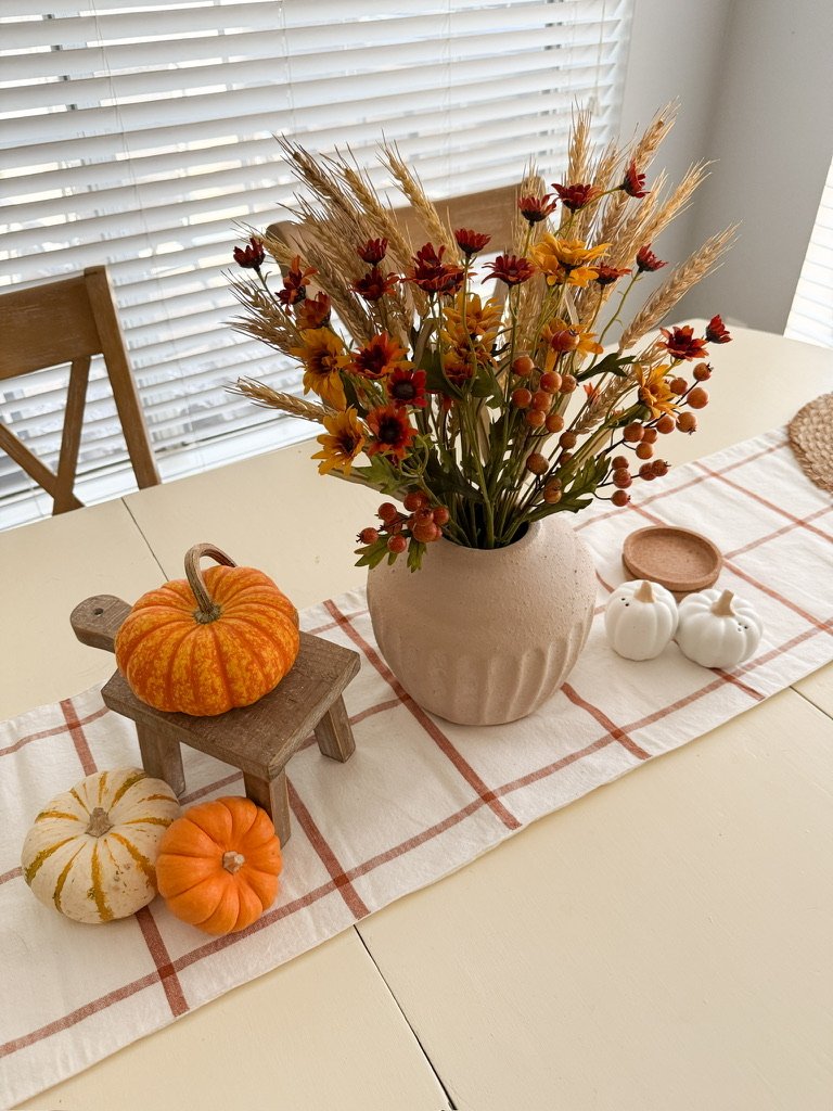 Fall kitchen table with a stone vase, white and orange table runner, mini pumpkins, and pumpkin salt and pepper shakers