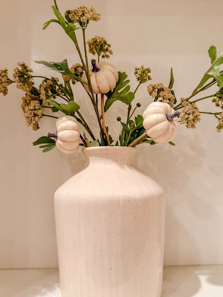 Neutral cream vase with greenery florals on a shelf with neutral white pumpkin picks. 