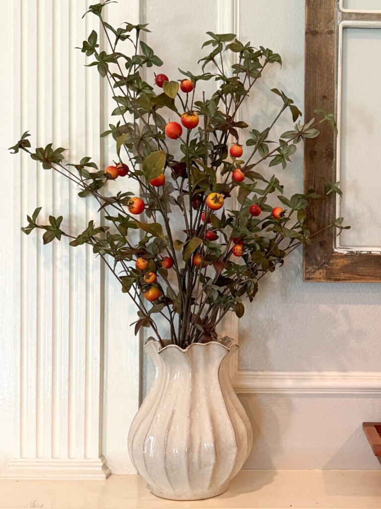 Cream fluted vase with greenery florals and orange berry stems on fireplace mantle.